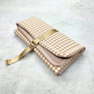 May include: A folded rectangular pouch featuring a tan and white gingham check pattern. The pouch is secured with a gold-colored fabric strap that loops through a matching gold metal D-ring, creating a functional closure for the textile accessory.