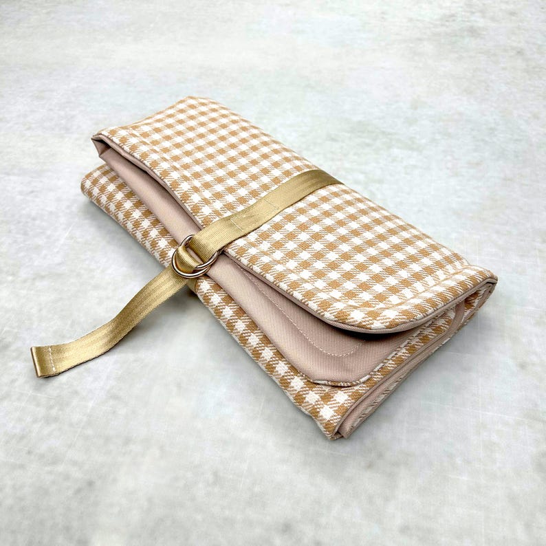May include: A folded rectangular pouch featuring a tan and white gingham check pattern. The pouch is secured with a gold-colored fabric strap that loops through a matching gold metal D-ring, creating a functional closure for the textile accessory.