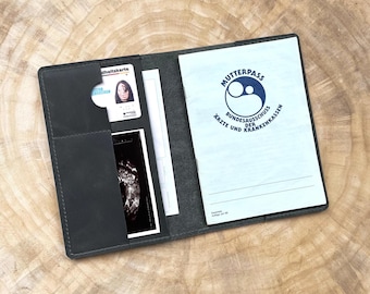 May include: A dark gray leather folder with a blue and white "Mutterpass" booklet inside. The booklet has a logo with a stylized image of a mother and child. The folder has a pocket for a health insurance card and a pocket for an ultrasound image.