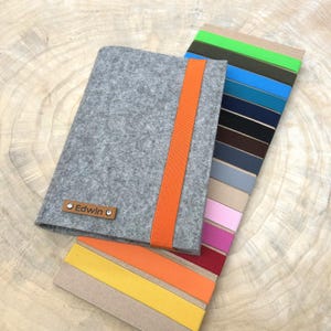 Può includere: Una copertina per taccuino in feltro grigio con una chiusura a fascia elastica verticale arancione brillante. Sul davanti è applicata una piccola etichetta rettangolare in pelle marrone con il nome Edwin in nero, fissata da due piccoli rivetti metallici argentati.