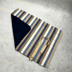May include: A rectangular fabric roll-up case featuring vertical stripes in shades of tan, white, and blue. The interior is lined with solid dark navy fabric. A beige strap with a gold-toned metal D-ring is attached to the exterior surface.