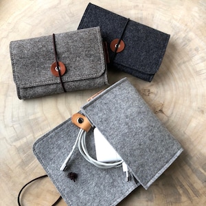 Può includere: Tre pochette in feltro grigio con dettagli in pelle marrone. Una pochette è aperta e contiene un cavo di ricarica bianco e un adattatore di alimentazione bianco.