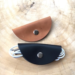 Könnte beinhalten: Zwei halbkreisförmige Kabelorganizer aus Leder mit silberfarbenen Metalldruckknöpfen. Ein Organizer ist hellbraun und der andere schwarz, wobei der schwarze Organizer ein aufgewickeltes weißes Ladekabel in seiner gefalteten Struktur hält.