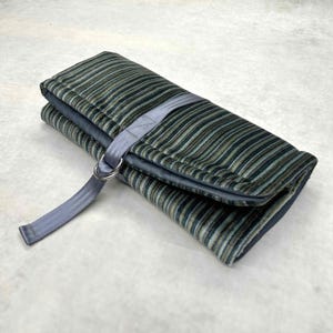 May include: A rectangular fabric roll-up case featuring a multi-colored striped pattern in shades of dark green, blue, and tan. The case is secured with a grey nylon strap and two silver-toned metal D-rings, keeping the folded textile closed.