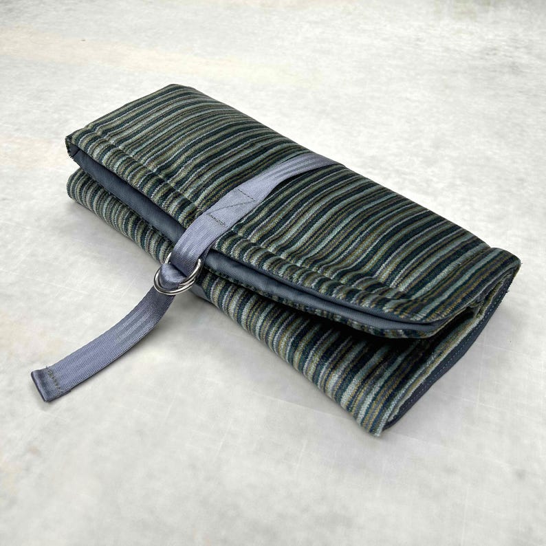 May include: A rectangular fabric roll-up case featuring a multi-colored striped pattern in shades of dark green, blue, and tan. The case is secured with a grey nylon strap and two silver-toned metal D-rings, keeping the folded textile closed.