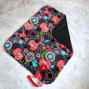 May include: A rectangular fabric mat featuring a vibrant pattern of red hearts, black and blue flowers, and green foliage on a dark background. The mat has a solid black underside and a red fabric strap with a metal ring attached to one corner.
