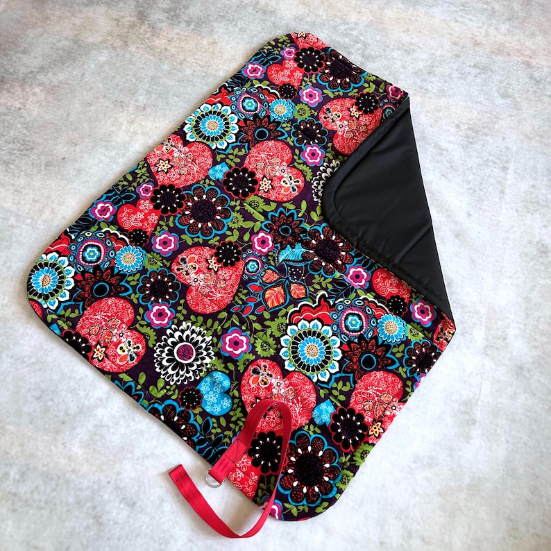 May include: A rectangular fabric mat featuring a vibrant pattern of red hearts, black and blue flowers, and green foliage on a dark background. The mat has a solid black underside and a red fabric strap with a metal ring attached to one corner.