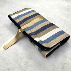 May include: A rectangular fabric roll-up case featuring a herringbone pattern in shades of navy blue, light blue, white, and tan. The case is secured with a tan nylon webbing strap that includes a metal D-ring closure on the exterior side.