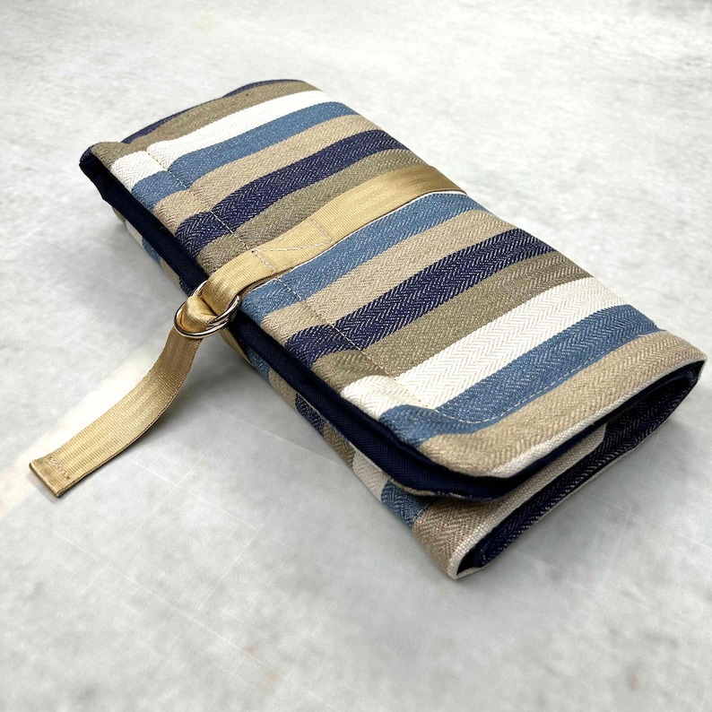 May include: A rectangular fabric roll-up case featuring a herringbone pattern in shades of navy blue, light blue, white, and tan. The case is secured with a tan nylon webbing strap that includes a metal D-ring closure on the exterior side.
