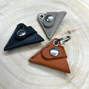 Könnte beinhalten: Drei Leder-Schlüsselanhänger in Dreiecksform. Die Schlüsselanhänger sind in Schwarz, Braun und Grau. Jeder Schlüsselanhänger hat einen silbernen Druckknopfverschluss.