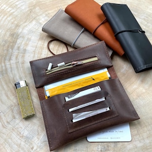 Può includere: Un astuccio per tabacco in pelle marrone con una tasca con cerniera, contenente sigarette e cartine. Un accendino dorato con un motivo strutturato è a sinistra. Diverse custodie in pelle arrotolata di vari colori sono sullo sfondo.
