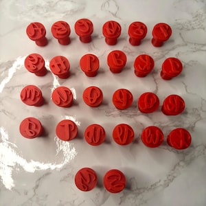 May include: A set of twenty-six red circular stamps, each featuring a unique raised letter of the alphabet in a cursive script font. The stamps are arranged in rows, with each piece having a cylindrical base and a flat top surface displaying the letter.
