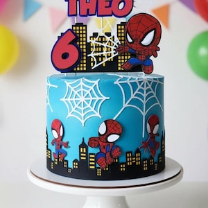 Può includere: Una torta di compleanno blu decorata con elementi a tema Spider-Man. La torta presenta un design a paesaggio urbano, ragnatele bianche e diverse figure di Spider-Man. Il cake topper recita "THEO" e il numero "6". La torta è su un alzatina bianca.