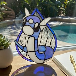 May include: Stained glass suncatcher featuring a blue and white cartoon character design. The suncatcher is in a teardrop shape and hangs from a chain. The image is set on a wooden surface with a succulent plant and notebook in the background.