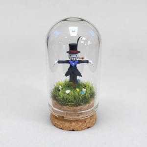 Puede incluir: Un terrario de vidrio con una figura de espantap&aacute;jaros caprichosa, con sombrero de copa negro y pipa. La figura se encuentra en un paisaje de hierba en miniatura con flores de colores. El terrario tiene una base de corcho.