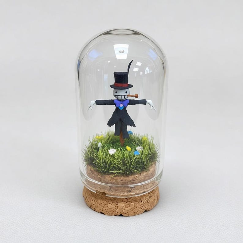 Puede incluir: Un terrario de vidrio con una figura de espantap&aacute;jaros caprichosa, con sombrero de copa negro y pipa. La figura se encuentra en un paisaje de hierba en miniatura con flores de colores. El terrario tiene una base de corcho.