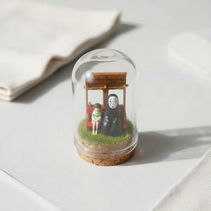 Puede incluir: Un terrario en miniatura con forma de c&uacute;pula de cristal que presenta dos figuras de una popular pel&iacute;cula de animaci&oacute;n. Las figuras est&aacute;n sentadas en un banco rojo bajo una estructura de madera, rodeadas de c&eacute;sped artificial. La base es de corcho.