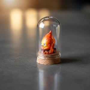 Puede incluir: Una peque&ntilde;a campana de cristal alberga una vibrante figura de personaje de llama naranja y amarilla. La figura se asienta sobre una base de corcho, con una peque&ntilde;a estructura de madera en su base. La c&uacute;pula de cristal es transparente y el fondo est&aacute; borroso.