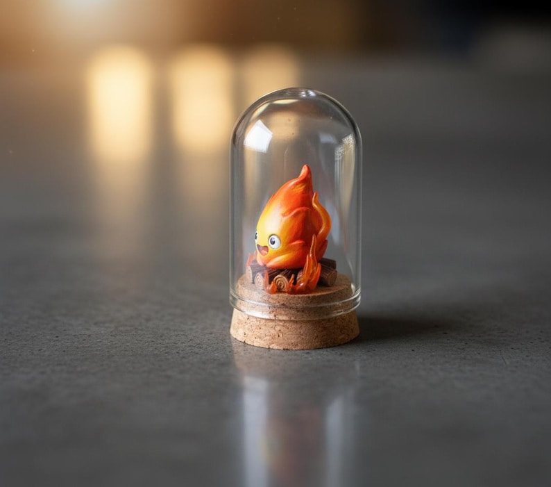 Puede incluir: Una peque&ntilde;a campana de cristal alberga una vibrante figura de personaje de llama naranja y amarilla. La figura se asienta sobre una base de corcho, con una peque&ntilde;a estructura de madera en su base. La c&uacute;pula de cristal es transparente y el fondo est&aacute; borroso.