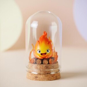 Puede incluir: Una figura de llama de dibujos animados con una cara sonriente, dentro de una c&uacute;pula de cristal. La llama es naranja y amarilla, sobre una base de corcho. El fondo es de un color neutro y suave.