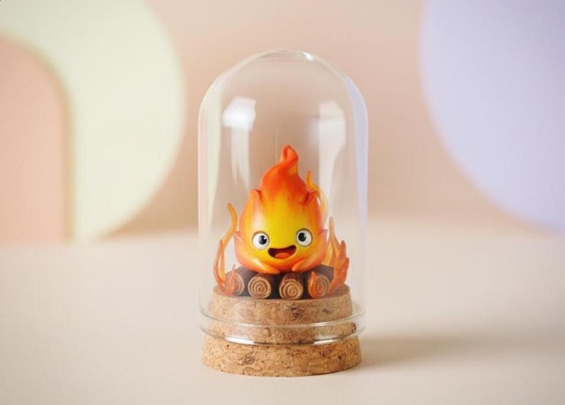 Puede incluir: Una figura de llama de dibujos animados con una cara sonriente, dentro de una c&uacute;pula de cristal. La llama es naranja y amarilla, sobre una base de corcho. El fondo es de un color neutro y suave.
