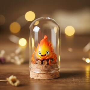 Puede incluir: Una figura caprichosa de un esp&iacute;ritu de fuego, con llamas naranjas y amarillas, encerrada en una c&uacute;pula de cristal. La base es de corcho y la figura tiene una cara sonriente. El fondo est&aacute; borroso con luces bokeh doradas y c&aacute;lidas.