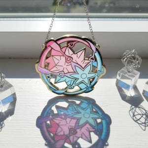 Può includere: Un elemento decorativo rotondo in vetro colorato con una cornice dorata. Il design presenta due elementi a forma di stella in rosa e blu, con dettagli a cuore e nastro. È appeso a una delicata catena.