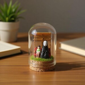 Puede incluir: Un diorama en miniatura dentro de una c&uacute;pula de cristal con dos figuras y una peque&ntilde;a estructura de madera. Una figura es una ni&ntilde;a con una camisa a rayas verdes y blancas, y la otra es una figura alta y negra con una m&aacute;scara blanca. La base es de corcho.