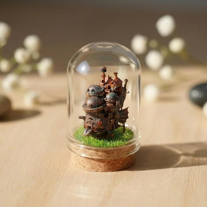 Puede incluir: Una escultura en miniatura de un castillo de estilo steampunk, encerrada en una c&uacute;pula de cristal. La escultura marr&oacute;n presenta detalles intrincados y se asienta sobre un lecho de c&eacute;sped artificial verde, con una base de corcho. La c&uacute;pula transparente se encuentra sobre un fondo borroso.