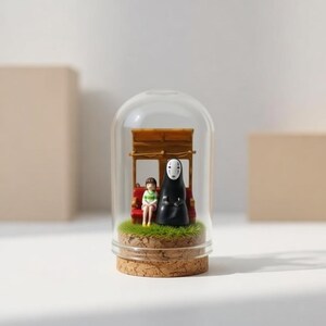 Puede incluir: Una c&uacute;pula de cristal decorativa con una escena en miniatura de una pel&iacute;cula. En el interior, una figura con una t&uacute;nica negra y un ni&ntilde;o est&aacute;n sentados en un banco rojo bajo una estructura de madera. La base es de corcho y c&eacute;sped artificial.