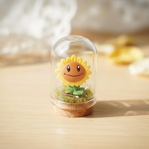 May include: A glass dome with a cork base contains a smiling sunflower figurine. The sunflower has yellow petals, a brown face, and green leaves. The base contains green moss.