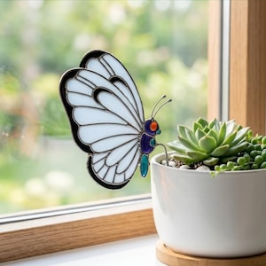 May include: A stained glass butterfly with white wings, black outlines, and colorful accents of orange, blue, and teal. The butterfly is positioned near a white potted succulent plant, creating a decorative scene.
