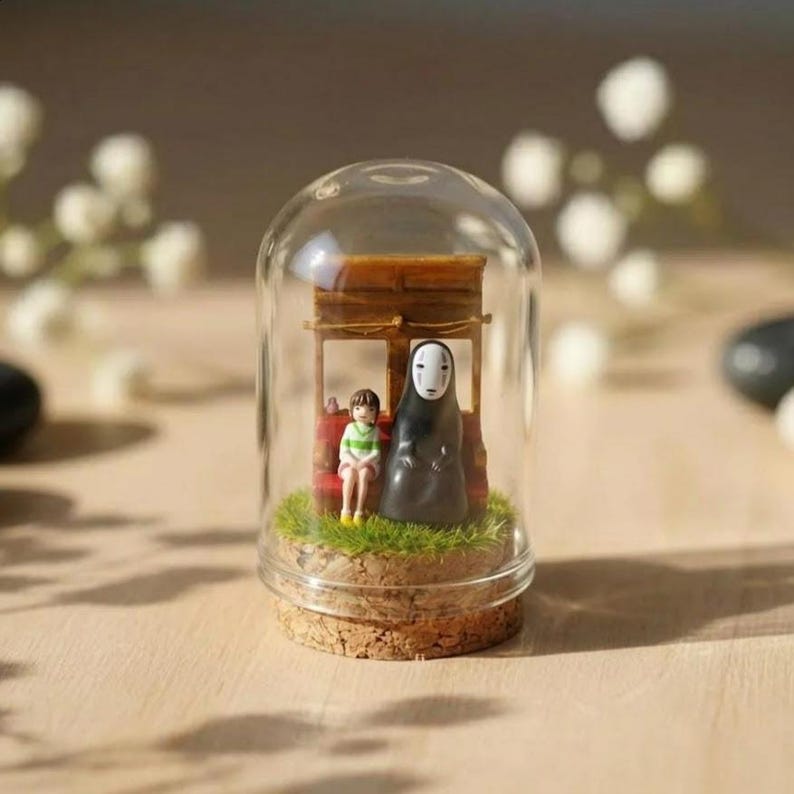 Puede incluir: Diorama en miniatura dentro de una c&uacute;pula de cristal con personajes de una pel&iacute;cula de animaci&oacute;n japonesa. La escena incluye una peque&ntilde;a estructura de madera, dos figuras y c&eacute;sped artificial, todo sobre una base de corcho.