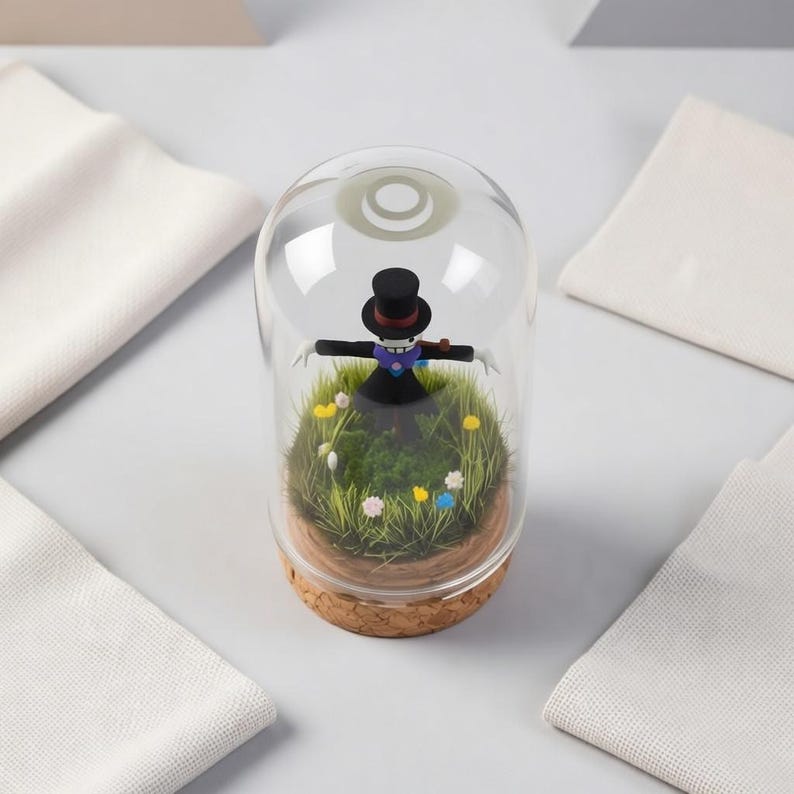 Puede incluir: Un terrario de vidrio decorativo con una figura en miniatura de un espantap&aacute;jaros con sombrero de copa y traje negro, sobre un lecho de musgo verde y flores coloridas. El terrario tiene una base de corcho.
