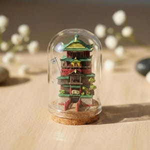 Op de afbeelding: Een miniatuurgebouw in Japanse stijl, voornamelijk rood en groen, gehuisvest in een glazen koepel. Het gebouw staat op een kurken voet. De koepel wordt gepresenteerd op een lichtgekleurd oppervlak. Het ontwerp is gedetailleerd.