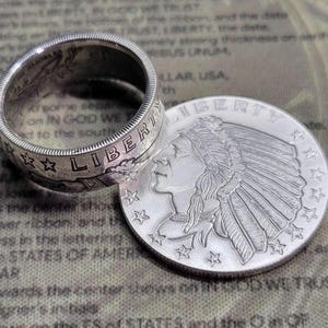 May include: A silver ring made from a US silver dollar coin featuring the words "LIBERTY" and "IN GOD WE TRUST". The ring is worn on a finger and the coin is facing the viewer. The coin is a US silver dollar with the image of a Native American woman wearing a headdress.