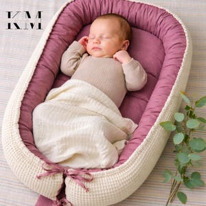 Może przedstawiać: Śpiące dziecko spoczywa w miękkim, owalnym gnieździe dla niemowląt. Gniazdo ma wnętrze w kolorze malwy, kremowy wafelkowy splot na zewnątrz i kremowy kocyk. Dziecko ma na sobie beżowe body. Gałązka zieleni znajduje się w tle.