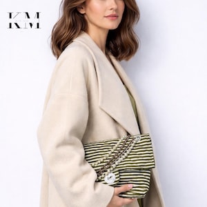 以下が含まれることがあります： ベージュのコートを着た女性が、パールとチェーンのストラップが付いた、緑と白のストライプのハンドバッグを持っています。 ハンドバッグはボタン留めです。 テキスト「KM WITH LOVE」が左上にあります。