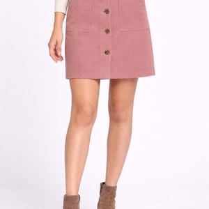 Op de afbeelding: Een stoffen rok in stoffig roze met twee voorzakken en knoopsluitingen. De rok wordt gecombineerd met bruine suède enkellaarsjes. Het model draagt een crèmekleurig shirt met lange mouwen.
