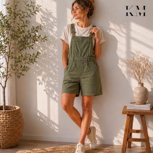 Può includere: Salopette corta verde oliva con spalline regolabili e tasca frontale. La salopette è abbinata a una t-shirt color crema e scarpe da ginnastica bianche. L'outfit è casual e confortevole, adatto all'uso quotidiano.