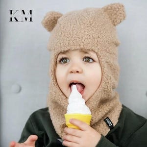 Könnte beinhalten: Ein Kind mit einem braunen Teddybär-Hut mit Ohrendetails, das ein weißes Eis in einer gelben Waffel isst. Der Hut hat einen Halsschutz und das Wort "talvi" an der Seite. Der Text "KM WITH LOVE" ist ebenfalls zu sehen.