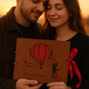 May include: A wooden photo album with a red hot air balloon design and the words "Our Love Story" is held by a couple. The album is tied with a red ribbon. The background is a warm sunset.