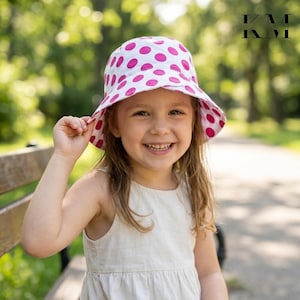 May include: A white bucket hat featuring a pattern of vibrant pink polka dots. The hat has a downward-sloping brim and a rounded crown. The fabric is lightweight and the design is consistent across the entire surface of the headwear.