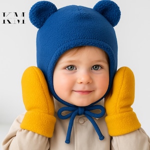 Peut inclure: Un enfant portant un bonnet d'ours en polaire bleu avec des liens bleus assortis et des mitaines en polaire jaune. Le chapeau a deux oreilles rondes sur le dessus. L'enfant porte un manteau beige.