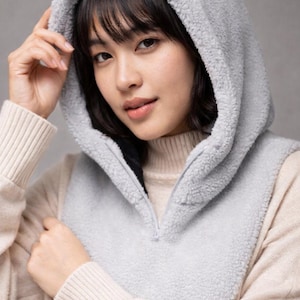 May include: A light grey hooded vest with a zip-up front. The vest is made of a soft, fuzzy material. The model is wearing a cream-coloured sweater underneath. The image also shows the text "KM TATKI".