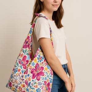 Peut inclure: Un sac fourre-tout floral avec un fond blanc et de grandes fleurs roses, jaunes et bleues. Le sac est porté à l'épaule. Le sac a une sangle florale assortie. Le logo de la marque est dans le coin supérieur gauche.