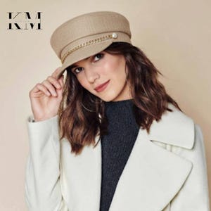 Pode incluir: Chapéu de marinheiro bege com detalhe de corrente dourada e um acento de pérola. O chapéu tem uma viseira estruturada e uma coroa arredondada. Acessório de moda.