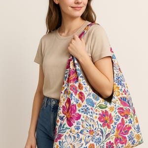 May include: A floral tote bag with a white background and large pink, blue, and orange flowers. The bag is held by a person wearing a beige t-shirt and blue jeans. The bag's strap has a floral pattern.