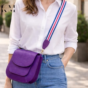 May include: A purple leather crossbody bag with a flap closure and a striped shoulder strap in red, white, and blue. The bag is worn with a white button-down shirt and blue jeans.