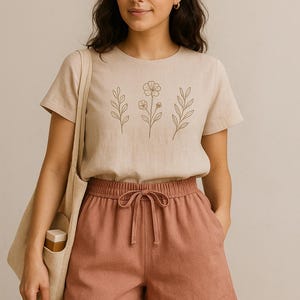 May include: A woman wearing a beige t-shirt with a floral design and rust-colored shorts. The t-shirt features a simple botanical illustration in a gold color. The shorts have a drawstring waist. A beige tote bag is over the shoulder.